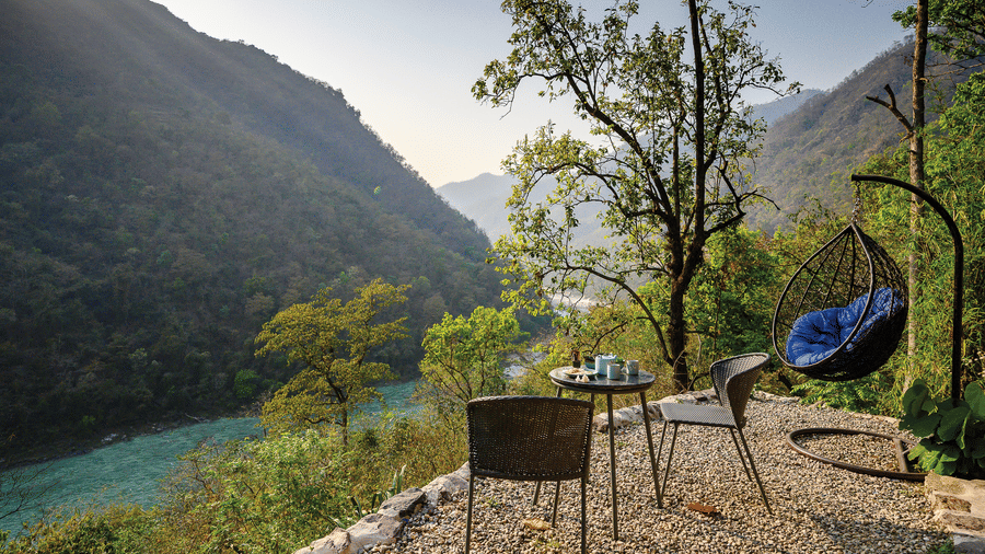 A scenic outdoor seating area featuring a small table with chairs and a purple hanging nest chair overlooking the turquoise waters of the river at Neemrana’s Glasshouse on The Ganges where guests can enjoy the tranquil mountain atmosphere and breathtaking 