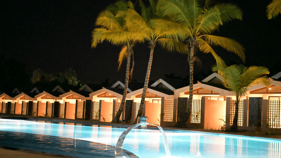 Symphony Palms Beach Resort And Spa's Havelock Pool Villas, showing multiple illuminated structures with pointed roofs and surrounding trees reflected in the calm surface of a swimming pool.