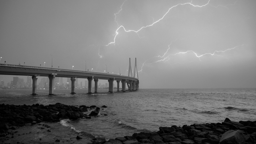 Black and white image of a long bridge extending over the sea with lightning striking in the cloudy sky above and rocky shoreline in the foreground.