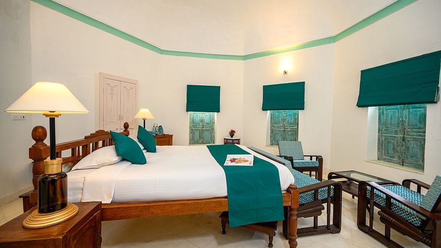 A bedroom with a wooden bed decorated with pillows and a runner, seating area, and a lamp on the bed side table. - Tijara Fort-Palace - 19th Century, Alwar