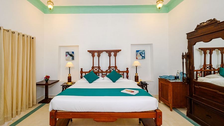 A bedroom with a wooden bed decorated with pillows and a bed runner, side lamps, framed art, and a wooden cabinet on the side. - Tijara Fort-Palace - 19th Century, Alwar