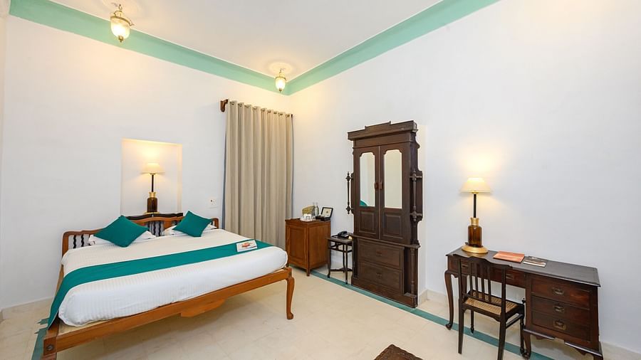 A room with a wooden bed, teal accents, a dark wood armoire, a desk with a chair, and two lamps, showcasing the room's simple elegance - Tijara Fort-Palace - 19th Century, Alwar.