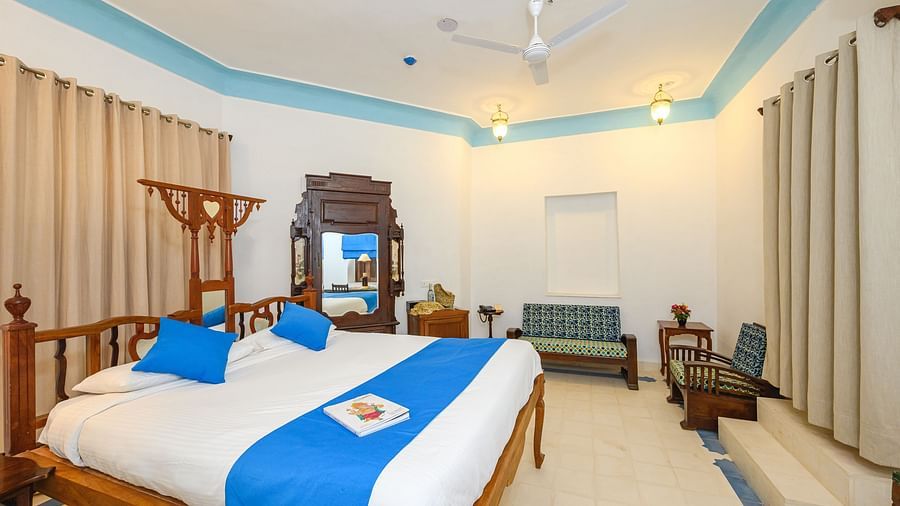 A bedroom with a wooden bed decorated with pillows and a bed runner, a seating area,and a cupboard in the background. - Tijara Fort-Palace - 19th Century, Alwar