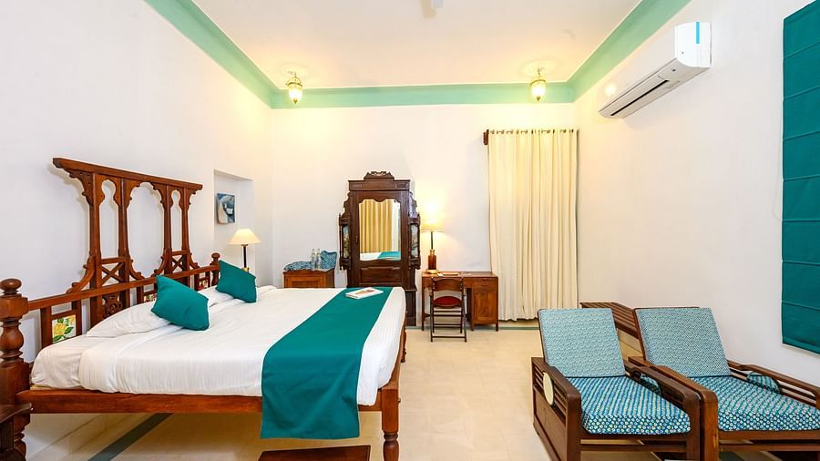 A spacious room with a wooden four-poster bed, teal accents, two armchairs, a desk, and an air conditioner, captured in a wide-angle shot - Tijara Fort-Palace - 19th Century, Alwar.