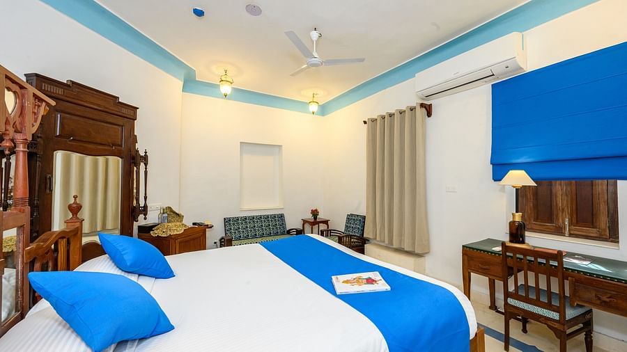 A bedroom with a wooden bed decorated with pillows and a bed runner, a seating area, a table with a chair, and a cupboard in the background. - Tijara Fort-Palace - 19th Century, Alwar