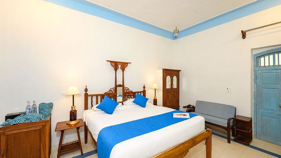 A bedroom with a wooden bed decorated with pillows and a runner and a door is on the other side. - Tijara Fort-Palace - 19th Century, Alwar