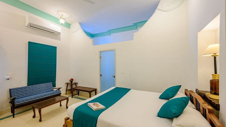 A room with a wooden bed, teal accents, a blue couch, two small tables, an air conditioner, and a unique ceiling design - Tijara Fort-Palace - 19th Century, Alwar.
