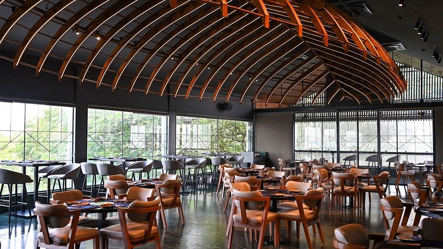 Contemporary restaurant dining room at Hotel Daspalla, Hyderabad featuring curved illuminated wooden ceiling, wooden chairs, dark tables, and floor-to-ceiling windows with garden views.