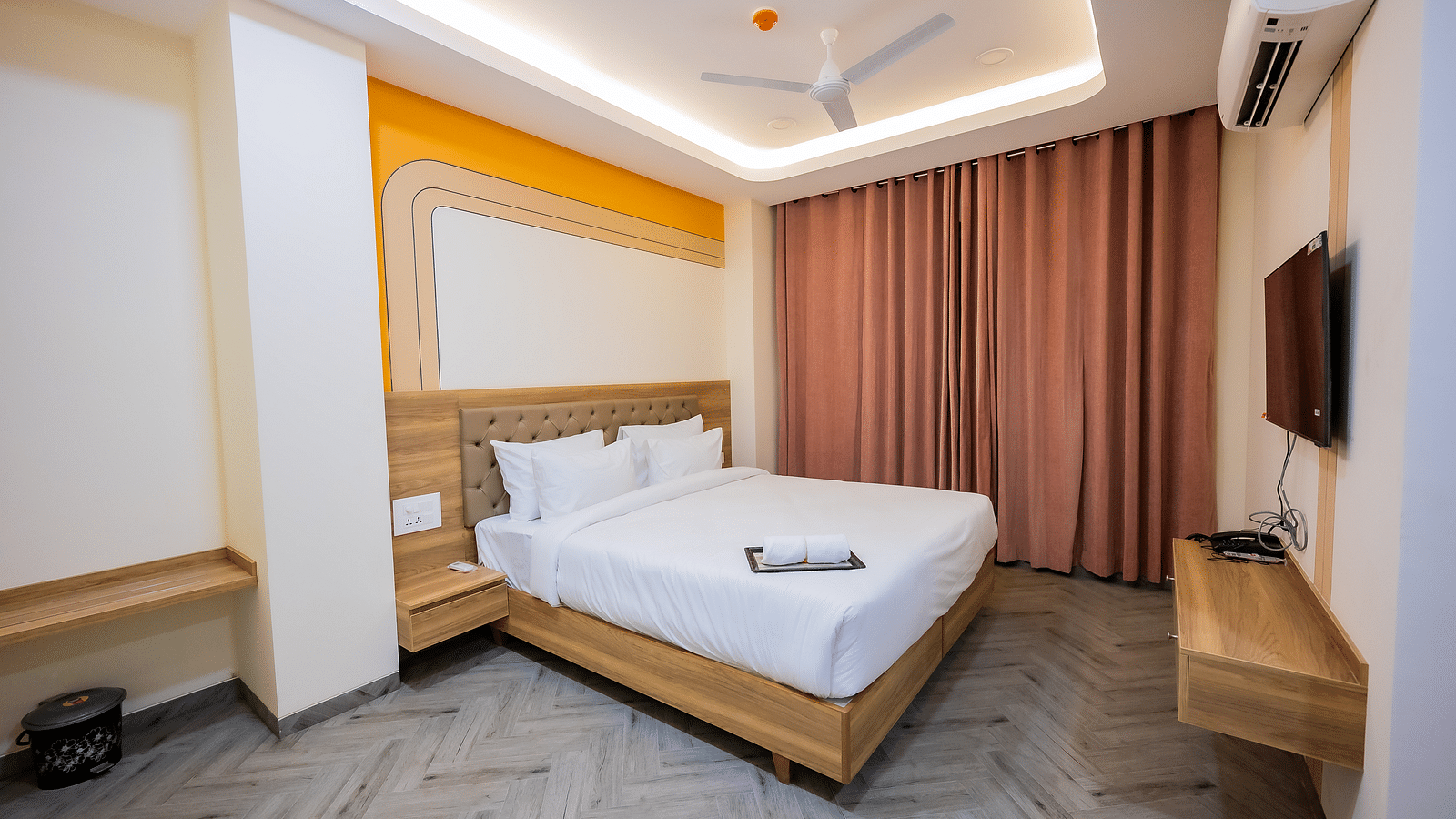 A cosy hotel room with soft lighting, a bed, curtains, and a TV mounted on the wall.