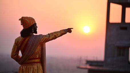 A person dressed as Shivaji Maharaj pointing to the sun with a building next to him.