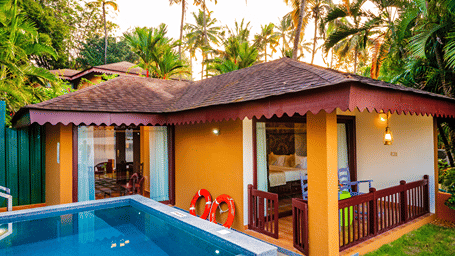 A villa exterior with a swimming pool in the foreground and lush greenery in the backdrop.