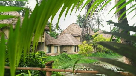 Row of Earth Villas at Vedic Village Spa Resort with mud walls and thatched roofs, facing a water body and enclosed by tall trees and thick foliage.