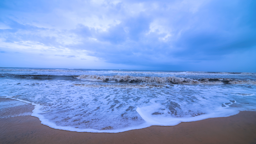 Large white waves crash onto a wide stretch of wet, dark sand under a heavy, blue-tinged, turbulent sky, suggesting an imminent storm.