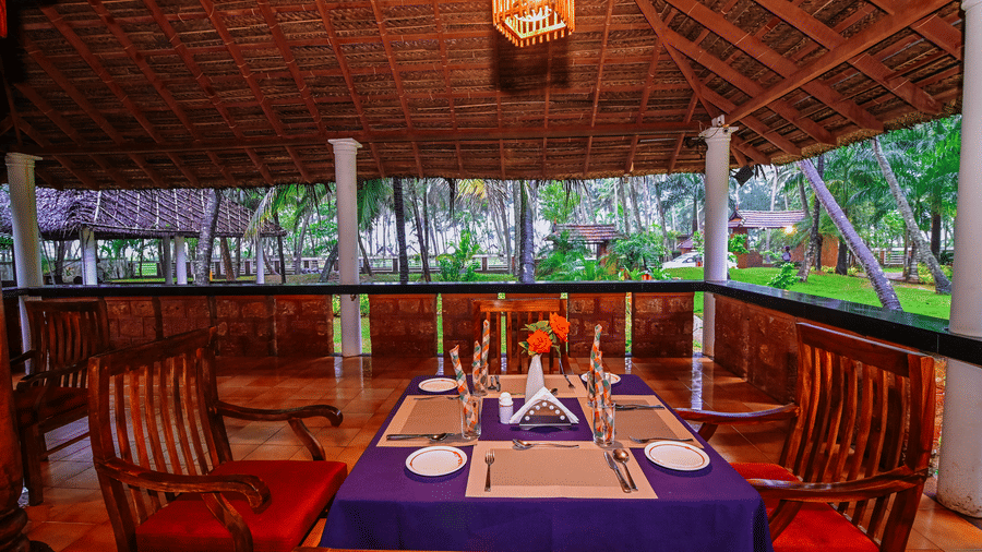 A dining table, set for two with a purple cloth, sits in an open-air pavilion with a thatched roof, surrounded by wooden chairs and columns.