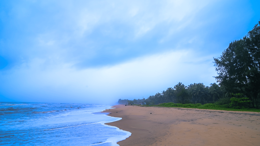 A deserted stretch of golden-brown sand meets the foaming turquoise-blue sea under a brooding, overcast sky, bordered by a line of dark green trees near Ayur On The Beach Nattika.