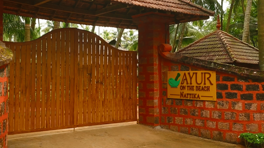A close up of a wooden gate leading to the Ayur On The Beach Nattika, with a thatched roof.