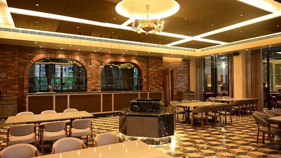 Dining room with tables, chairs, patterned floor, and textured walls, lit by ceiling lights and a chandelier, with a counter in the background.