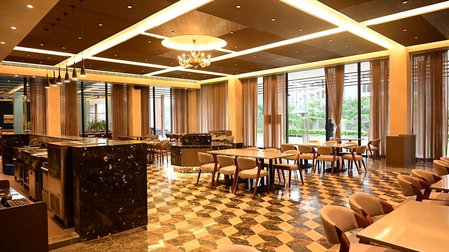 Dining area with tables, chairs, patterned floor, and large windows, lit by ceiling lights and a chandelier.