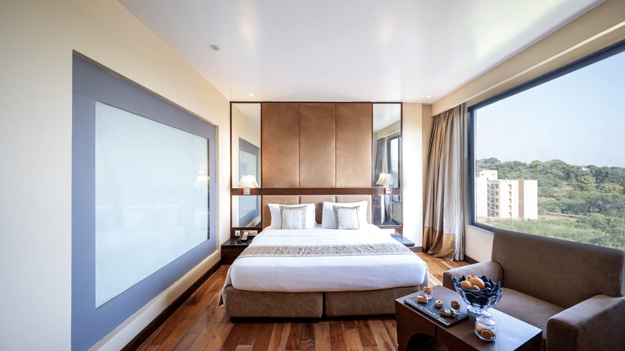 Premium Room at Essentia Luxury Resort & Spa, Udaipur featuring a plush bed by a larg window with the view of trees and a seating space near the window