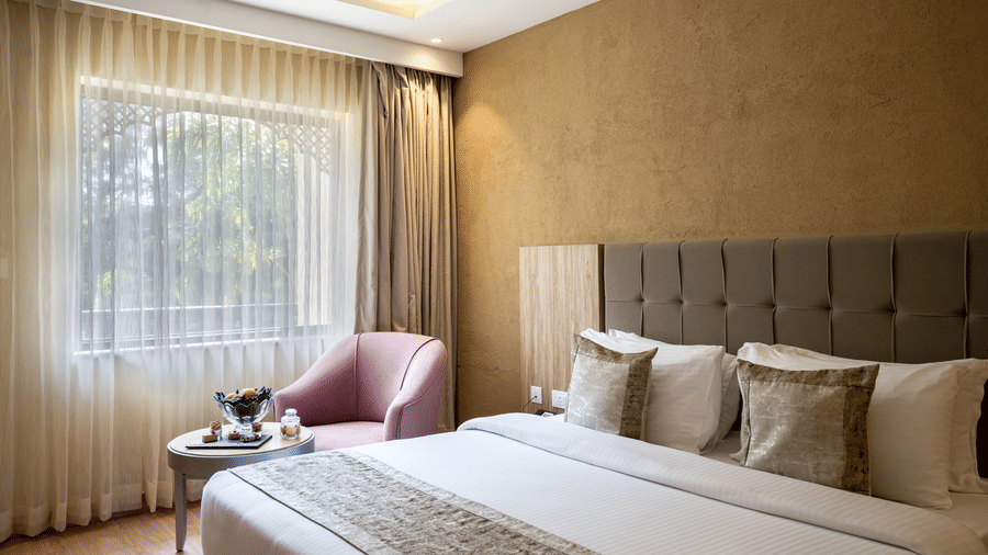 Standard Room at Essentia Luxury Resort & Spa, Udaipur featuring a plush bed with pillows, cushions and a bed runner next to a seating space by the window with sheer curtains