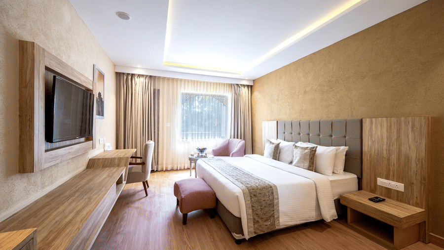 Standard Room at Essentia Luxury Resort & Spa, one of the best places to stay in Udaipur, featuring a plush bed with ottoman facing the TV on the wall over a running desk with a chair by the window with a seating area