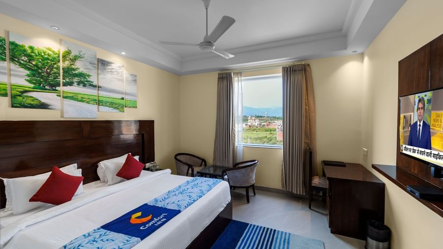 Modern furnished room with a double bed featuring a ceiling fan, artwork, chairs by the window, and flat-screen TV on the side wall at Inde Reva Resorts Rishhikesh.
