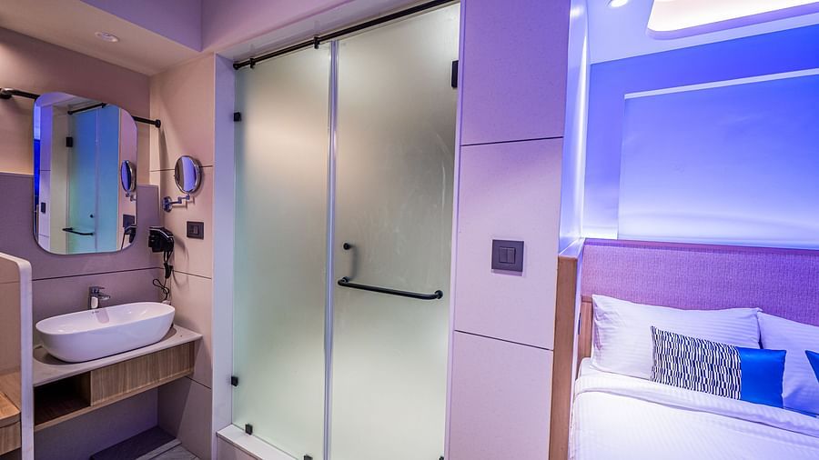 A washbasin below a wall-mounted mirror with a tinted door beside it, and a bed placed next to it at Indie Stays Mumbai International Airport.