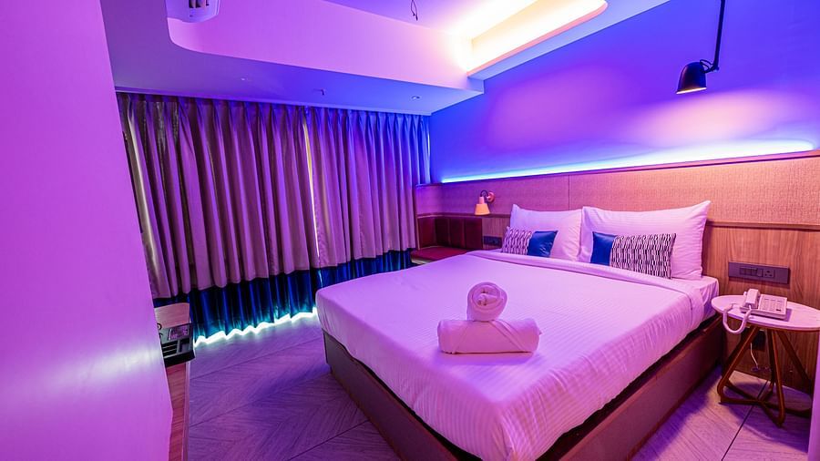 A spacious room at Indie Stays Mumbai International Airport features a large bed with neatly rolled towels, a window with closed curtains, and the room lit with cool lighting.