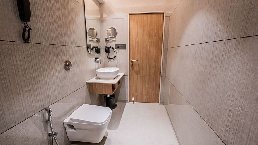 A spacious washroom at Indie Stays Mumbai International Airport, featuring a wooden door, a wall-mounted mirror above the basin, a western toilet, and a shower area.