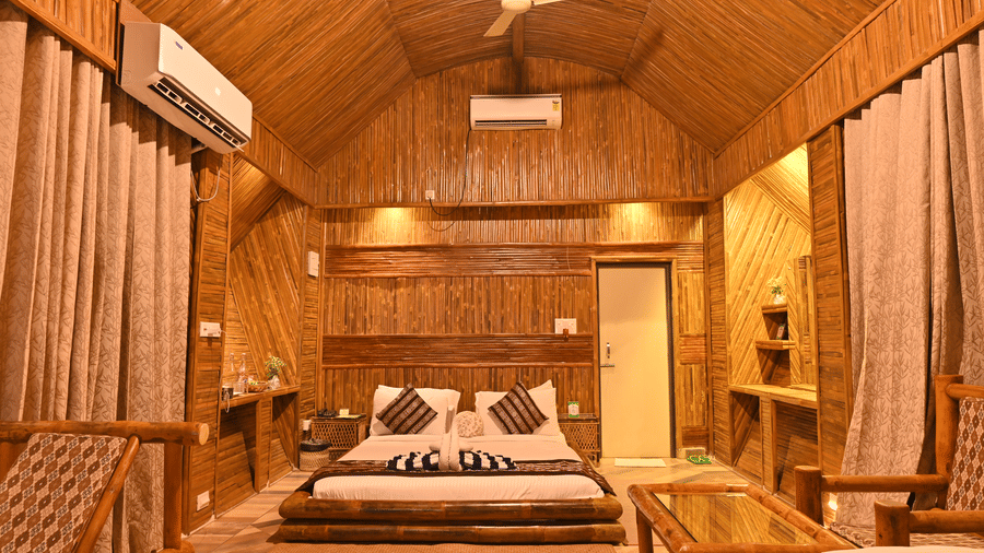 Bamboo cottage bedroom with a bed dressed in patterned linen, a wooden ceiling fan, long curtains and warm wooden panelled walls throughout.