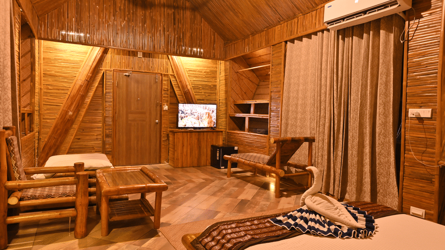 Bamboo cottage interior with wooden panelled walls, a wall-mounted television, wooden seating, a bed and a sloped wooden ceiling above it.