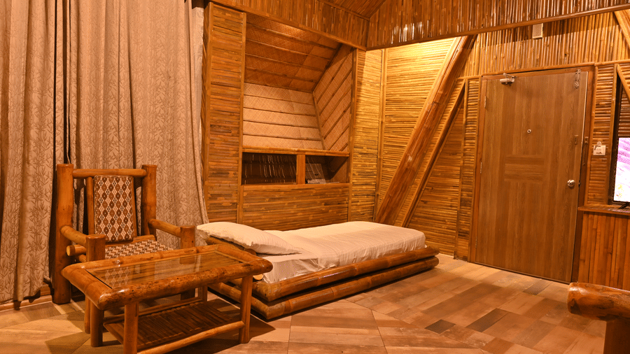 Bamboo cottage interior with wooden panelled walls, a sloped thatched ceiling, a wooden bench, a bed and long beige curtains along the window.