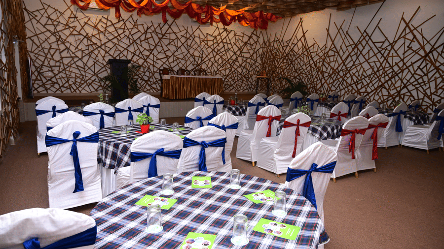 An indoor view of an event space at Pipul Odi Art Museum Resort, set up for a small gathering with tables covered in checked cloths and chairs draped with white and blue covers, under a bamboo-lined ceiling.