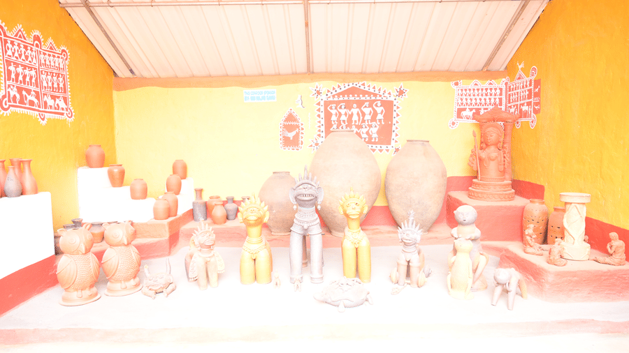 A brightly lit indoor exhibit hall showcasing a wide array of terracotta pottery and tribal figurines arranged around a central seating area, with yellow and white walls, at Pipul Odi Art Museum Resort.