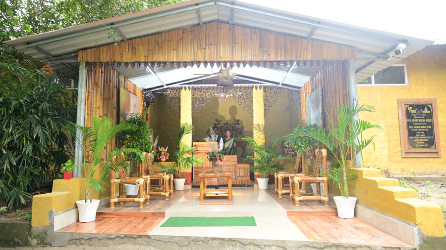 The open-fronted reception area of Pipul Odi Art Museum Resort, constructed with bamboo walls and a corrugated metal roof, featuring wooden furniture, potted palm plants, and a person seated behind a desk in the interior.