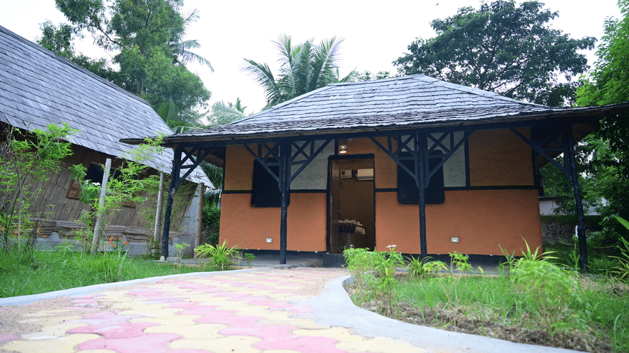 An exterior view of one of the bamboo or tribal cottages at Pipul Odi Art Museum Resort, featuring an orange wall and a dark, thatched roof, set amongst a green lawn and paved pathway.