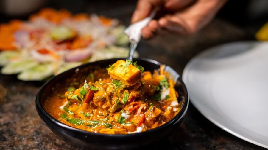 A bowl of curry is being served at Pravasa Stay, Shirdi, with a decorative vegetable platter in the background.