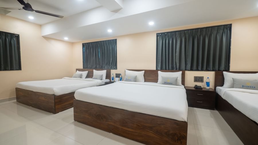 A large room at Pravasa Stay, Shirdi, contains three wooden platform beds with white linen and dark curtains.