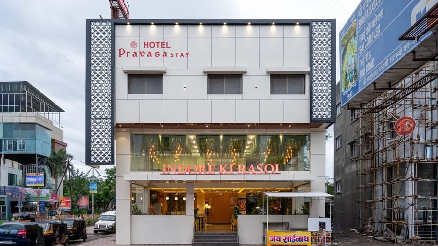 The main facade of Pravasa Stay, Shirdi, a white three-storey building, has a ground floor entrance and windows.