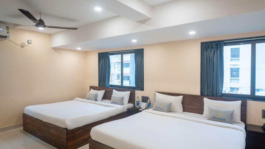 A room at Pravasa Stay, Shirdi, has two wooden beds, two large windows, and a ceiling fan installed.