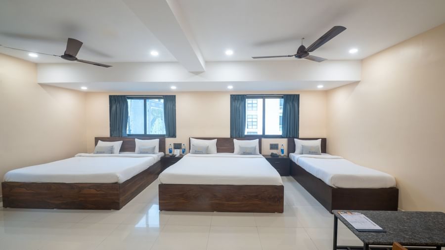 A large room at Pravasa Stay, Shirdi, contains three beds, two ceiling fans, and multiple windows.