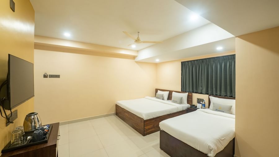 A room at Pravasa Stay, Shirdi, is equipped with two beds, a wall-mounted television, and dark curtains over a window.