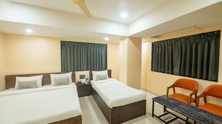 A room at Pravasa Stay, Shirdi, has two beds, two orange chairs, a small table, and a ceiling fan.