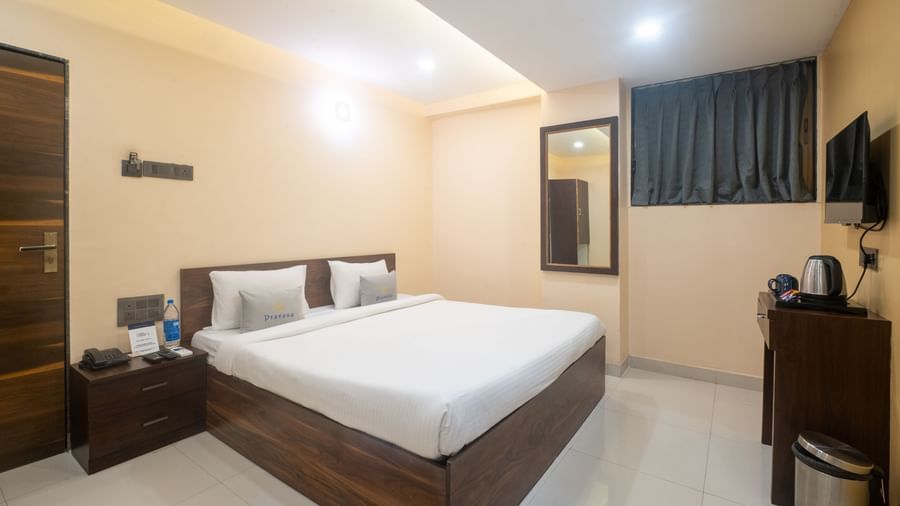 A room at Pravasa Stay, Shirdi, is furnished with a wooden bed, a bedside table, and a wall-mounted mirror.