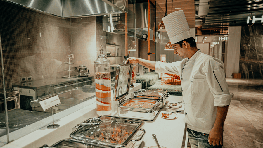 A chef in a white uniform prepares food behind a counter in a professional kitchen. There are different dishes in metal containers on the counter.