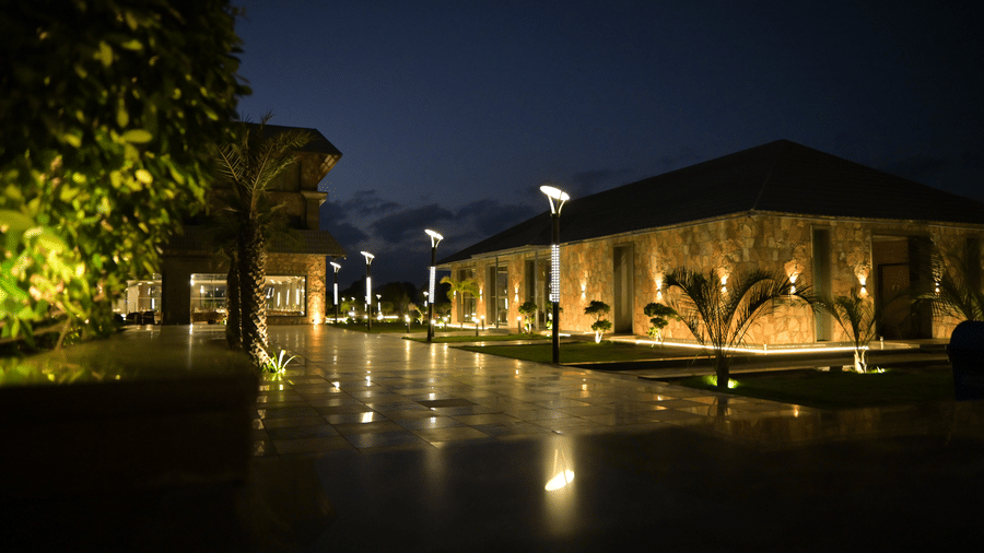 A resort building is brightly lit at night, with lights reflecting on the wet ground. The manicured lawn and building are surrounded by trees.