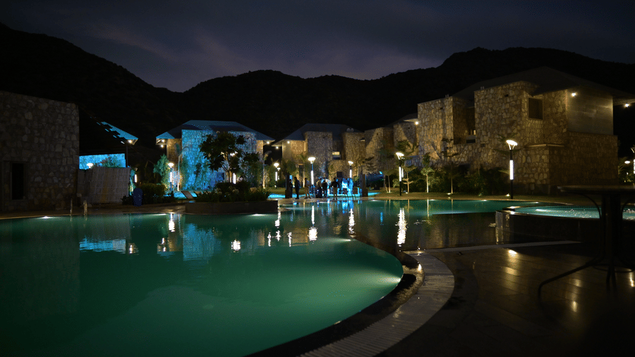 A large, curving swimming pool and a deck are lit up at night, reflecting the warm lights of the resort buildings.