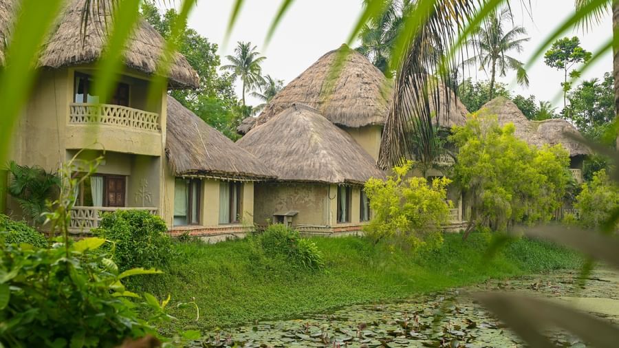 Thatched-roof Earth Villas at Vedic Village Spa Resort overlooking a water body, with lush green vegetation lining the perimeter of the natural surroundings.