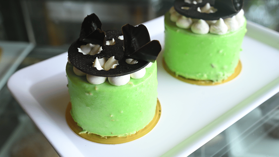 Two green mousse cakes topped with dark chocolate decorations served on a white platter at Vedic Village Spa Resort displayed in a glass showcase in an artistic presentation.