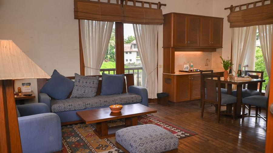 Living area of the Lake View Villa at Vedic Village Spa Resort with blue couches, a large rug, wooden furnishings, a sink near the dining table, and natural light entering through large windows.